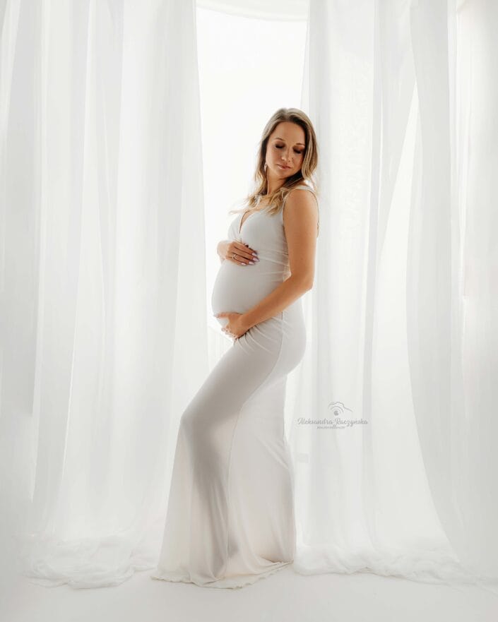 Przyszła mama w białej sukience trzyma dłonie na brzuchu w otoczeniu delikatnych zasłon – sesja ciążowa Warszawa